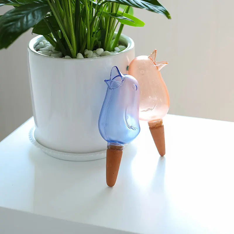 Self-watering bird-shaped plant stakes in a white indoor plant pot with green leaves.