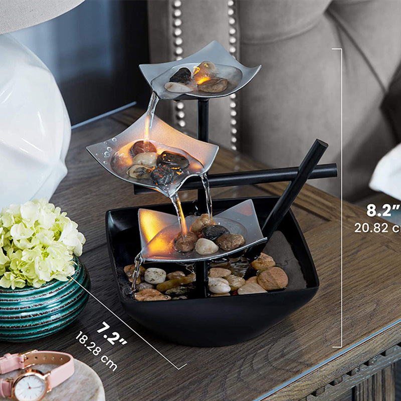 Decorative water fountain with rocks on a wooden surface, surrounded by flowers and a watch.